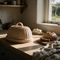 Woven rattan bread cloche on a pale oak kitchen bench beside a cut sourdough loaf in afternoon light.