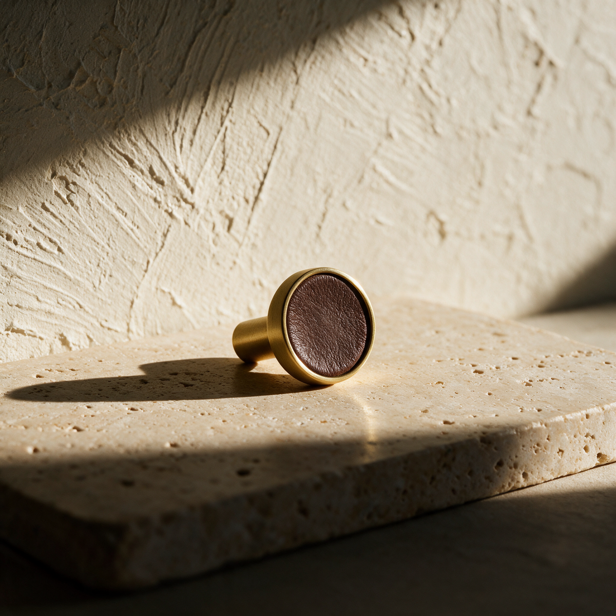A single brass and leather knob lying on its side on a travertine slab.