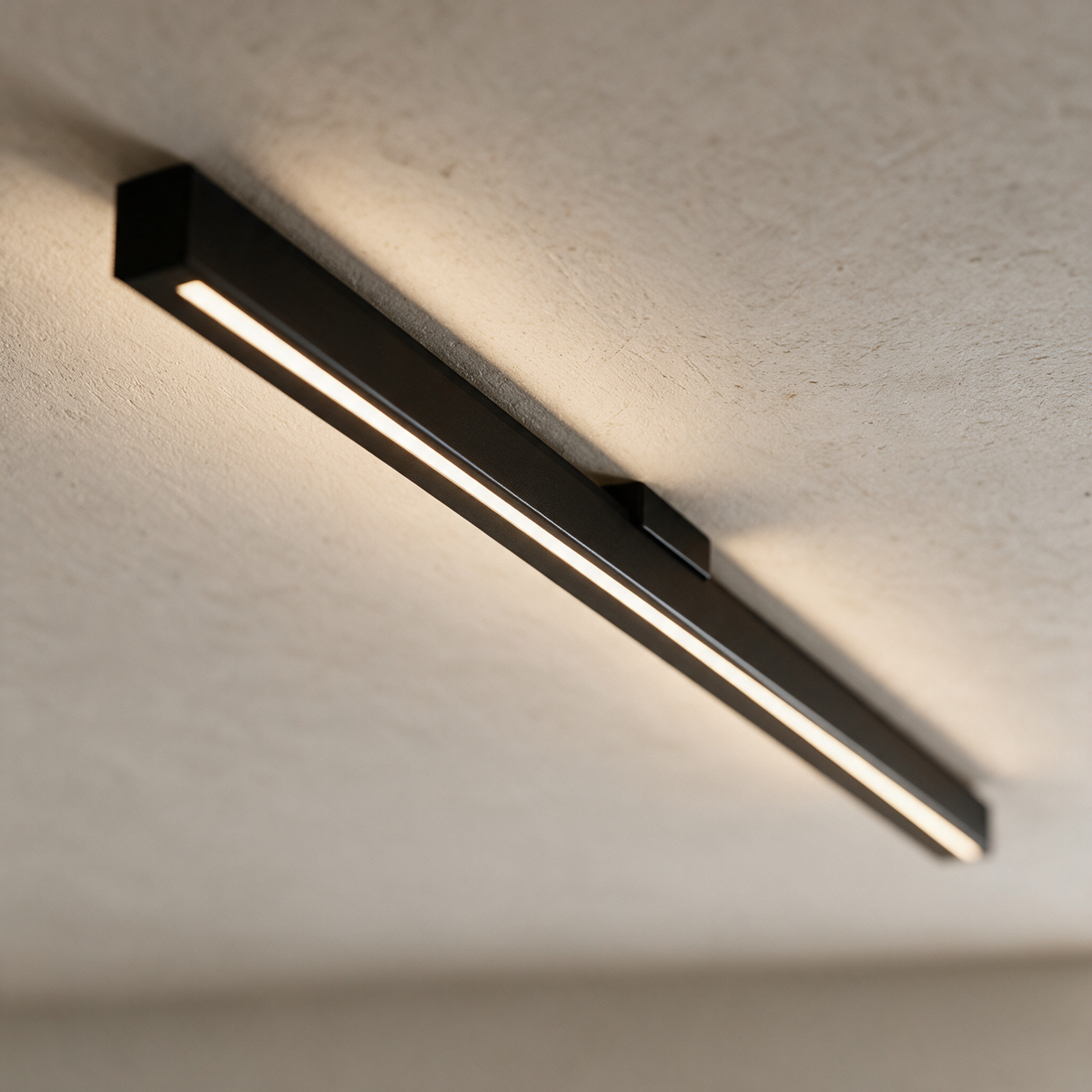 Close view of a slim matte black linear ceiling light glowing against a plaster ceiling