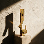The gold folded mixer alone on a travertine plinth against a textured plaster wall.