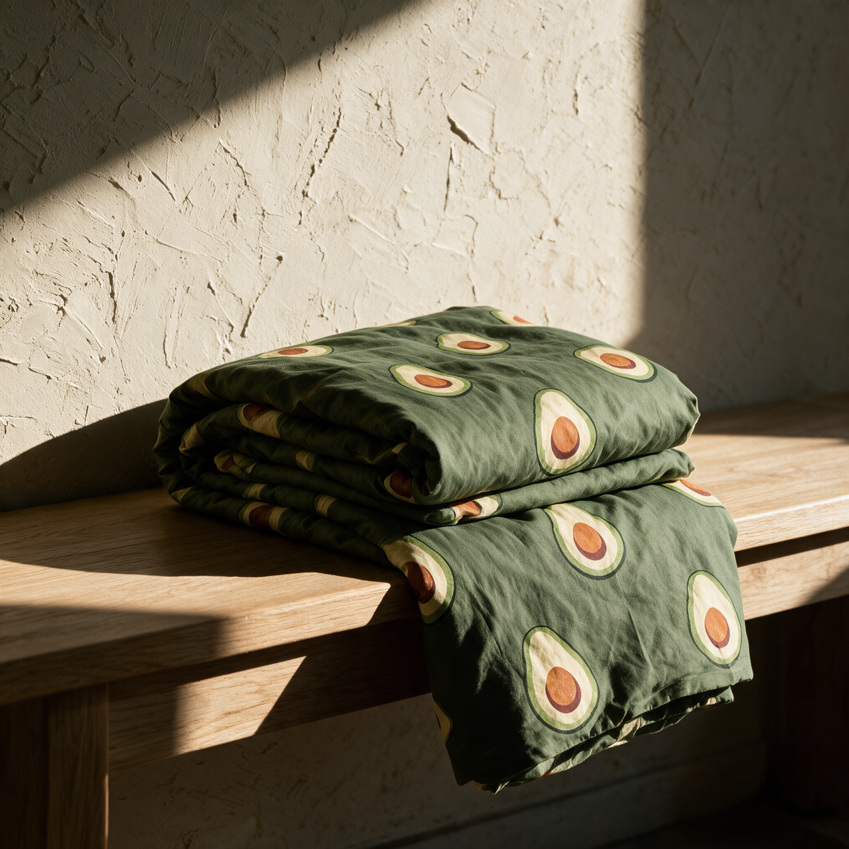 The avocado print quilt cover folded in a loose stack on an oak bench against a plaster wall.