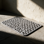 The dry shower mat alone on a concrete floor in hard raking light showing its drainage holes.