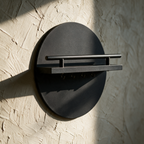 A round black wall key holder with a shelf, alone on a textured plaster wall in raking light.