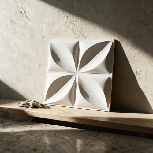 A single square white panel with a raised radial petal relief resting upright on an oak bench against a plaster wall.