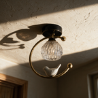 Unlit brass ring light with fluted glass and ceramic bird seen from below against a textured plaster ceiling in raking afternoon sun.
