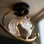 Macro close-up of the small white ceramic bird's feet resting on the brass ring with the ribbed glass shade behind.