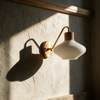 Unlit brass wall sconce with a white opal shade casting a long curved shadow on a sunlit plaster wall.
