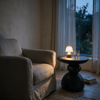 Matte black stacked pebble side table beside a linen armchair with a lit lamp at dusk
