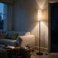 Slim black floor lamp with a cream cylinder shade lit beside a linen sofa at dusk.