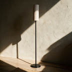 Unlit slim floor lamp on a pale oak floor casting a long line of shadow on plaster.