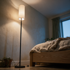 Low view of the lit floor lamp rising in a bedroom corner beside a bed.