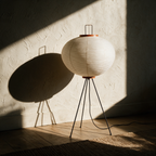 Unlit paper globe tripod lamp on a pale oak floor in raking afternoon sun.