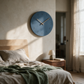 Matte blue wall clock with wooden hands above a bed in soft morning light