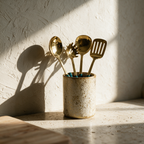 The full utensil set in its crock standing alone on a stone counter in hard raking light.