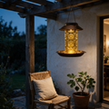 Bronze bird feeder lantern glowing amber under a pergola at dusk.