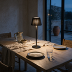 A cordless table lamp with a fluted shade and round black base lit on a dining table at dusk.