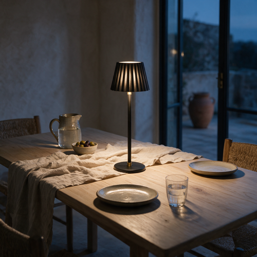 A cordless table lamp with a fluted shade and round black base lit on a dining table at dusk.