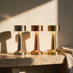 Three compact cordless table lamps in silver copper and brass on a travertine ledge