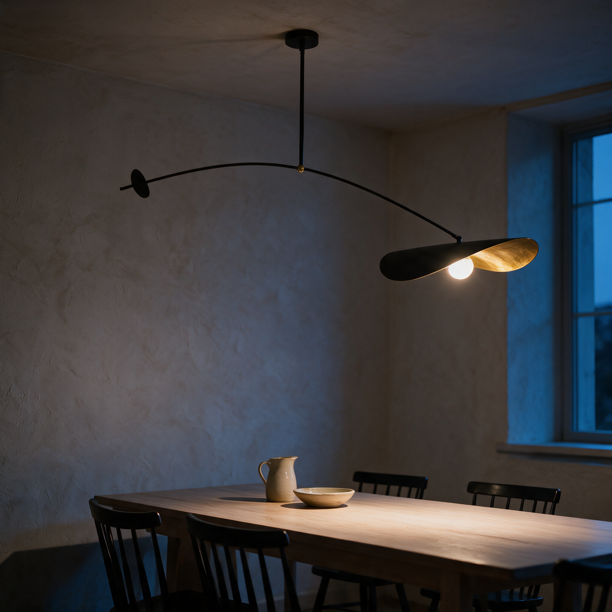 Balanced mobile pendant light with one leaf shade and a disc counterweight above a dining table at dusk